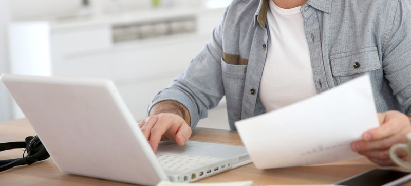 Man on white laptop with report in hand - stock image